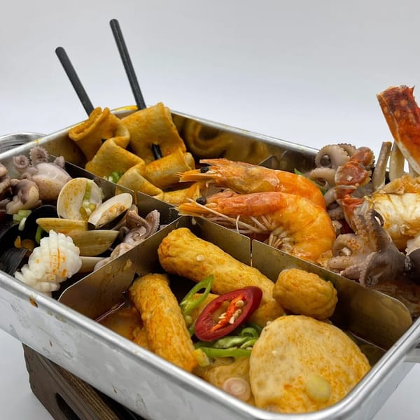 A metal tray with dividers holds various Korean seafood and fish cakes, including shrimp, octopus, and clams.