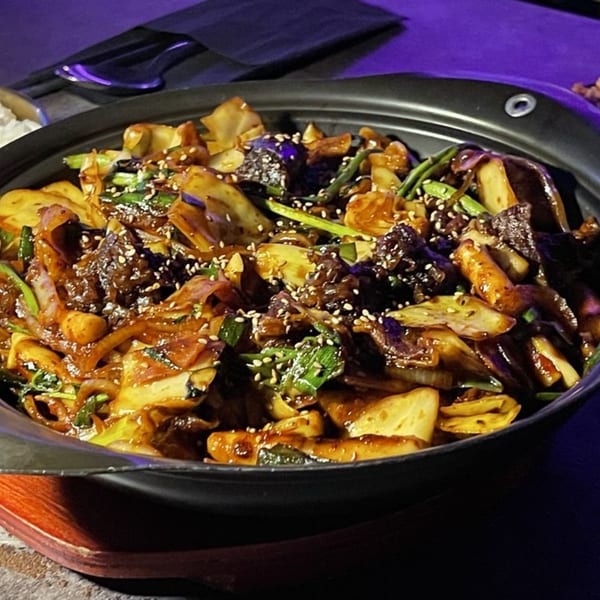 A close-up of a dark pot filled with a Korean stir-fry dish, garnished with sesame seeds.