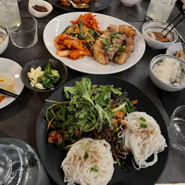 A table laden with various Korean dishes, including sliced pork belly, kimchi, noodles, and rice.